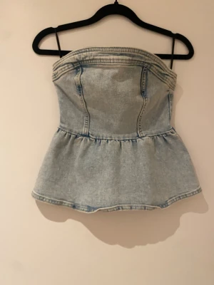 Ljusblå axelbandslös jeanskorsett - Trendig axelbandslös korsettopp i ljusblå denim med markerade sömmar och volang nedtill. 