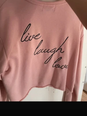 Rosa croppad topp från NA-KD - Säljer en ljusrosa croppad topp från Zorannah for NA-KD med svart broderad text 'live laugh love' på framsidan. Toppen har långa ärmar och en avslappnad passform, perfekt för en chill look.