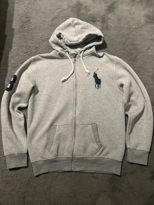 Grå zip hoodie från Polo Ralph Lauren - Snygg grå zip hoodie från Polo Ralph Lauren med stor mörkblå polospelare broderad på bröstet och nummerdetalj på ärmen. Hoodien har dragsko i huvan, kängurufickor och ribbade muddar. Tillverkad i mjuk bomullsblandning för extra komfort.
