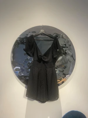 Svart kortklänning med puffärm - Supersöt svart kortklänning med puffärmar och v-ringning. Klänningen har en knyt-detalj framtill och är gjord i ett lätt, prickigt tyg som ger en luftig känsla. Perfekt för dig som vill ha en trendig och bekväm look.