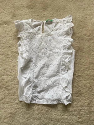 Vit ärmlös blus med volangdetaljer - Söt vit blus i bomull med broderade hålmönster och volangdetaljer längs axlarna. Ärmlös modell med rund halsringning och knapp i nacken. Perfekt för en fräsch och somrig look.