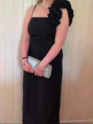 Svart balklänning  - Snygg svart långklänning med oneshoulder-design och stor volangdetalj på axeln. Klänningen har smal axelrem på ena sidan och är figurnära med elegant fall. Perfekt för dig som vill sticka ut på festen.