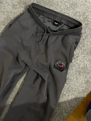 Gråa mjukisbyxor från Zavetti Canada - Snygga grå mjukisbyxor från Zavetti Canada med broderad logga och patch på ena benet. Byxorna har dragsko i midjan, muddar vid bensluten och praktiska fickor med dragkedja. Stora textdetaljer längs benen för en trendig look.