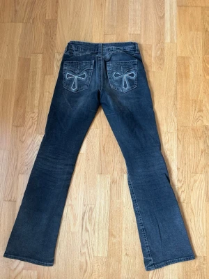 Svarta bootcut jeans med brodyr - Snygga svarta jeans med bootcut-modell och broderade detaljer i form av rosetter på bakfickorna. Jeansen har klassisk femficksdesign och subtila slitningar för en trendig look. Perfekta för dig som gillar en unik touch på dina jeans.