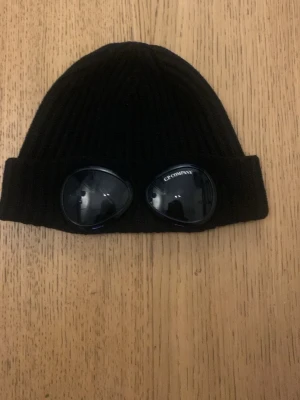 Svart goggle-mössa från C.P. Company - Säljer en svart ribbstickad mössa från C.P. Company med ikoniska goggle-linser på uppviket. Mössan är tillverkad i mjukt stickat material.