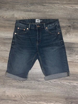 Mörkblå jeansshorts från 157 - Snygga mörkblå jeansshorts från 157 med klassisk femficksdesign och uppvikta benslut. Shortsen har normal passform och stängs med knapp och dragkedja. Perfekta för sommardagar och enkla att matcha med olika stilar.