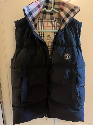 Svart väst från Burberry - Stilren svart dunväst från Burberry med klassiskt rutigt foder och hög krage. Västen har två fickor framtill, dragkedja och ett Burberry-emblem på bröstet. Perfekt för lager på lager och ger en lyxig känsla till din outfit.