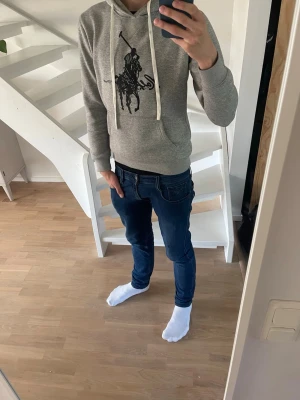 Grå hoodie från Polo Ralph Lauren - Snygg grå hoodie från Polo Ralph Lauren med stor svart logga och text på bröstet. Hoodien har huva med vita dragsnören och en stor magficka framtill. Tillverkad i mjukt sweatshirtmaterial som är perfekt för chill dagar. Tröjan har ett litet litet hål i ärmen som knappt syns.