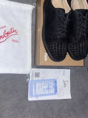Christian Louboutin svarta sneakers - Svarta sneakers från Christian Louboutin i mocka med svarta nitar på tån och svarta skosnören. Klassisk låg modell med exklusiv känsla och ikonisk design. Perfekta för dig som vill sticka ut med en lyxig och edgy stil.