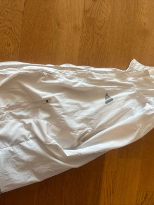 Vit vindjacka från Adidas - Snygg vit vindjacka från Adidas med dragkedja och diskret logga på bröstet. Jackan är tillverkad i lätt och tunt material, perfekt för blåsiga dagar. Praktisk ficka med dragkedja på sidan. Enkel och stilren design som passar till det mesta.
