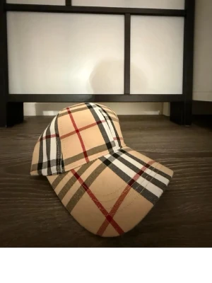 Beige keps med rutigt mönster - Snygg beige keps med klassiskt rutigt mönster i svart, vitt och rött. Kepsen har böjd skärm och justerbart spänne bak. Perfekt accessoar för en trendig streetstyle-look.