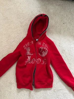 Röd hoodie Aim For The Moon XS - Snygg röd hoodie med dragkedja från Aim For The Moon. Tryck på framsidan med text och motiv av en björn och måne. Mjuk insida och huva med svart kant. Perfekt för dig som gillar streetwear och vill sticka ut. den har använts en gång och har inga skavanker eller trasiga delar. Den är i ett jättefint skick.