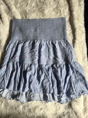 Ljusblå volangkjol med smock - Supersöt ljusblå kortkjol med smock i midjan och volanger. Kjolen har små blommiga detaljer i rosa och vitt, vilket ger en romantisk vibe. Perfekt för vår och sommar, och tillverkad i ett lätt bomullstyg som känns luftigt.