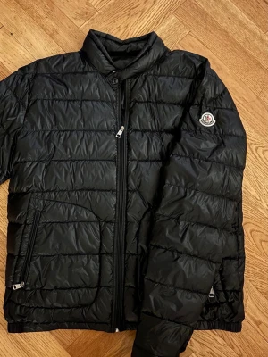Moncler acorus svart - Stilren svart pufferjacka från Moncler med diskret logga på ärmen. Jackan har ståkrage, dragkedja framtill och två fickor med dragkedja. Perfekt för kalla vinterdagar och ger en clean look. Kvitto från vongreven, samt äkthetsbevis via Monclers hemsida! Hör av dig vid funderingar