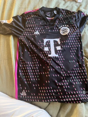Bayern München Musiala matchtröja - Säljer en svart och rosa Bayern München matchtröja från Adidas med Musiala och nummer 42 på ryggen. Tröjan har klubbmärke, sponsorlogga och färgglada detaljer framtill. Perfekt för fotbollsfans som vill sticka ut på läktaren eller planen.