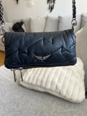 Svart quiltad axelväska från Zadig & Voltaire - Snygg svart axelväska i quiltat läder från Zadig & Voltaire. Väskan har ett coolt silverfärgat vingemblem framtill, kedjerem med läderdetaljer och dragkedja upptill. Perfekt storlek för det viktigaste och ger en edgy vibe till din outfit. Det finns lite rost på och defekter på kedjan (se bilder)