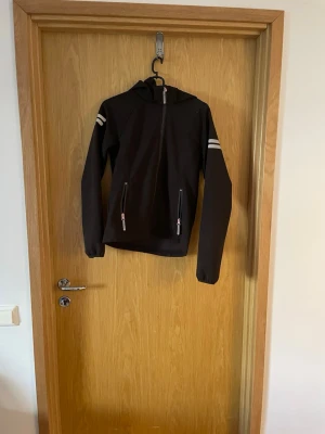 Svart vindjacka med vita ränder - Svart vindjacka med huva och två dragkedjeförsedda fickor framtill. Jackan har vita dubbla ränder på båda ärmarna och en smal passform. Perfekt för blåsiga dagar och enkel att matcha med olika outfits.