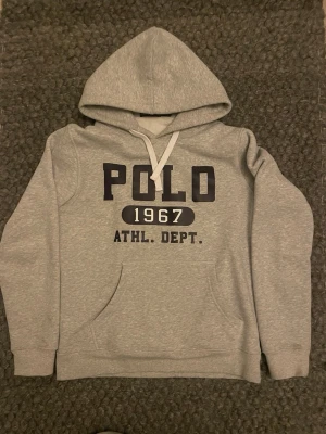 Grå hoodie från Polo Ralph Lauren - Snygg grå hoodie från Polo Ralph Lauren med stor huva och magficka. Tryck framtill med texten 'POLO 1967 ATHL. DEPT.' i svart och vitt. Klassisk modell med ribbade muddar och dragsko i huvan. Perfekt för en avslappnad streetwear-look. OBSS FINNS FLÄCKAR PÅ ARMEN OCH PASSAR SOM EN S 