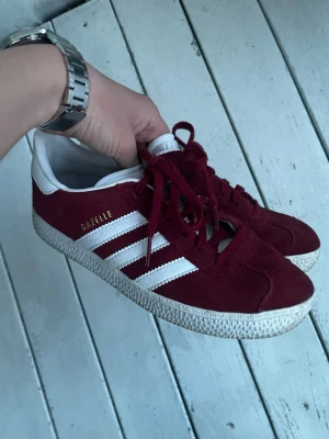 Adidas Gazelle vinröda sneakers - Säljer ett par klassiska Adidas Gazelle sneakers i vinrött mocka med vita detaljer och de ikoniska tre ränderna på sidan. Skorna har vit sula och vit hälflik med Adidas-logga. Perfekta för dig som gillar retrostil och streetwear. Väldigt fint skick och nästan aldrig använda. 1300kr+ nypris! Skriv vid frågor eller funderingar🙌✅