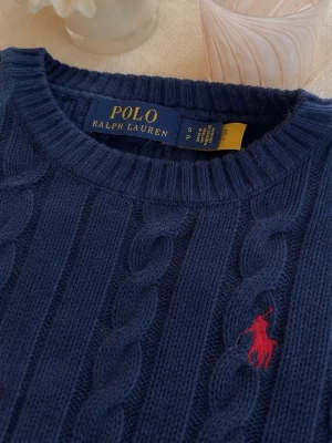 Mörkblå kabelstickad tröja Polo Ralph Lauren - Kabelstickad mörkblå tröja från Polo Ralph Lauren med rund halsringning och klassisk röd broderad logga på bröstet. Tröjan har ribbade muddar och är tillverkad i ett mjukt stickat material. Perfekt för en stilren och tidlös look. Skicka för mer info som bilder o frågor!!!                                Den är i bra skick förutom möjligtvis lite nopprig men inget som syns när man har den.            Pris går o diskutera vid snabb affär!!