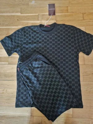 Svart set från Gucci med GG-mönster - Snyggt svart set från Gucci med klassiskt GG-mönster över hela både t-shirt och shorts. T-shirten har rund hals och korta ärmar, shortsen har resår i midjan. Perfekt för dig som gillar exklusiv och trendig stil.