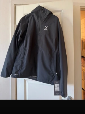 Svart regnjacka från Haglöfs - Snygg svart regnjacka från Haglöfs med dragkedja framtill och två sidofickor. Jackan har minimalistisk design, hög krage och är tillverkad i vattentätt material. Perfekt för regniga dagar och outdoor-äventyr.