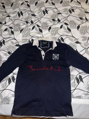 Mörkblå långärmad skjorta Bondelid - Mörkblå långärmad skjorta från Bondelid med vit krage och knappslå. Röd broderad Bondelid-logga på bröstet och en patch med emblem. Skjortan har klassisk rugbyinspirerad stil och är i bomull.