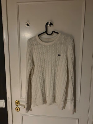 Vit kabelstickad tröja Gant - Kabelstickad vit tröja från Gant med rund halsringning och diskret logga på bröstet. Tröjan har långa ärmar och ribbade muddar vid ärmslut och nederkant. Perfekt för dig som gillar klassisk och stilren design.
