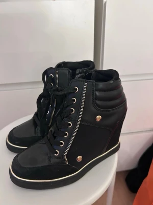 Svarta wedge sneakers med gulddetaljer - Snygga svarta sneakers med klack och guldfärgade detaljer, Skorna har snörning framtill och vadderad kant runt ankeln. Använda få gånger 