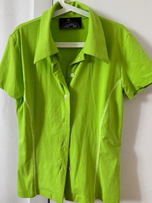 Limegrön kortärmad skjorta - Snygg limegrön skjorta med korta ärmar och klassisk krage. Skjortan har vita knappar framtill och dekorativa sömmar längs sidorna. Perfekt för dig som vill sticka ut med färg! Materialet känns mjukt och bekvämt.