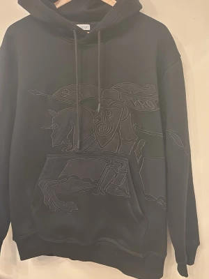 Svart hoodie från Burberry med brodyr - Svart hoodie från Burberry med stor broderad design framtill som föreställer en riddare på häst. Klassisk huva med dragsko och stor känguruficka. Tillverkad i mjukt material som känns lyxigt och bekvämt. Perfekt för dig som gillar streetwear med exklusiv känsla. PASSAR L 