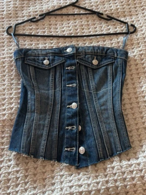Jeans korsettopp från Zara - Säljer en snygg jeans korsettopp från Zara i mörkblå denim med knappar framtill och två fejkade bröstfickor. Toppen är axelbandslös och har coola sömdetaljer som ger en edgy look. Perfekt att styla med höga byxor eller kjol!