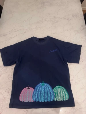 Mörkblå t-shirt med färgglada pumpor - Cool mörkblå t-shirt med tre stora färgglada pumpor broderade längst ner på ryggen. Pumporna är i gult, orange och rosa med detaljer i blått och grönt. Perfekt för dig som gillar unika och lekfulla plagg.