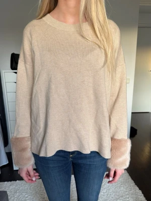 Beige stickad tröja med fuskpäls - Beige stickad tröja med rund hals och vida ärmar. Ärmsluten har breda detaljer i matchande beige fuskpäls.