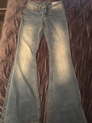 Blå bootcut jeans från Vera Moda  - Säljer ett par blå bootcut jeans från Vera Moda med klassisk femficksdesign och ljusa slitningar på framsidan. Jeansen har en låg midja och är tillverkade i bomullsdenim. Perfekta för dig som gillar retrostil och breda ben.