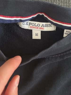 Mörkblå topp från U.S. Polo Assn. - Mörkblå topp från U.S. Polo Assn. med klassisk broderad logga på bröstet. Toppens insida av kragen har en snygg randig detalj i rött, vitt och blått. Tillverkad i mjuk bomull som känns skön mot huden.