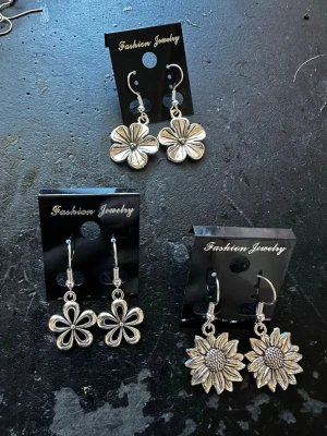 Tre par blommiga örhängen i silverfärg - Säljer tre par örhängen i silverfärg. Alla har blommotiv: ett par med solrosor, ett par med stiliserade blommor och ett par med klassiska blomblad. Alla är i metall och har krokar för enkel användning. Perfekta för dig som gillar blommiga accessoarer.