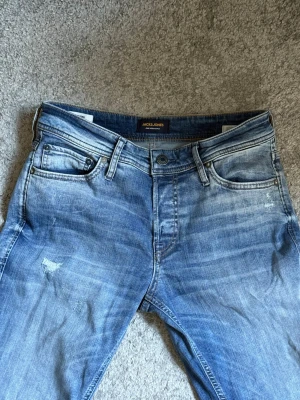 Jack&Jones jeans  - Säljer ett par blå jeans från Jack & Jones med snygga slitningar och ljus tvätt. Storlek 30x32, slim/glenn passform. Klassisk femficksmodell med dragkedja och knapp. Jeansen har en smal passform och är tillverkade i bomull med lite stretch för extra komfort.