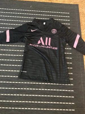 Svart PSG träningströja Nike S - Svart långärmad träningströja från Nike för PSG med rosa detaljer och logga. Tröjan har halv dragkedja, klubbmärke på bröstet och tryckt sponsor. Tillverkad i lätt och ventilerande funktionsmaterial, perfekt för fotbollsträning.