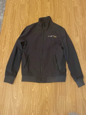 Svart vindjacka Sail Racing Gore-Tex - Snygg svart vindjacka från Sail Racing med Gore-Tex-material. Jackan har hög krage, hel dragkedja framtill och två snedställda fickor med dragkedja. Diskret logga på bröstet i vitt och gult. Perfekt för blåsiga dagar och sportig stil.