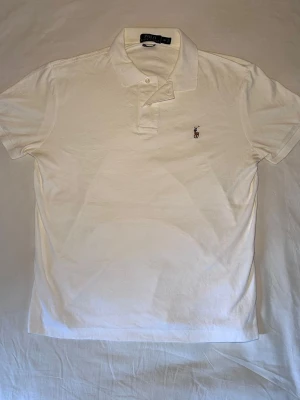 Ralph Lauren | Piké | M - Klassisk vit pikétröja från Polo Ralph Lauren med broderad logga på bröstet. Tröjan har korta ärmar, krage och knappar framtill. Tillverkad i mjuk bomull för en bekväm känsla och stilren look. Mått: 63 Längd 50 Bredd