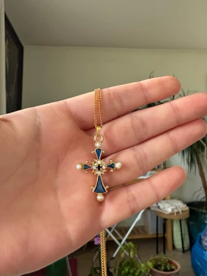 Guldigt halsband med blått kors - Snyggt halsband med ett guldfärgat kors som har blå emaljdetaljer och vita pärlor i varje ände. Kedjan är tunn och elegant, och korset har en unik vintagekänsla med dekorativa detaljer i mitten.