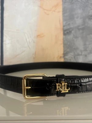 Svart bälte från Lauren Ralph Lauren - Stilrent svart bälte från Ralph Lauren med krokodilpräglat mönster och guldfärgat RL-spänne. Bältet har en klassisk design med elegant metallspänne och passar perfekt till jeans eller kostymbyxor. Inte använt. Plasten sitter kvar på RLL loggan. Köptes från Zalando. Strl 85.
