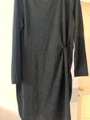 Svart glittrig långärmad klänning - Snygg svart klänning med glittriga detaljer och lång ärm. Modellen är rak med knyt i midjan som ger en fin siluett. Perfekt för dig som gillar en stilren look med lite extra glitter.