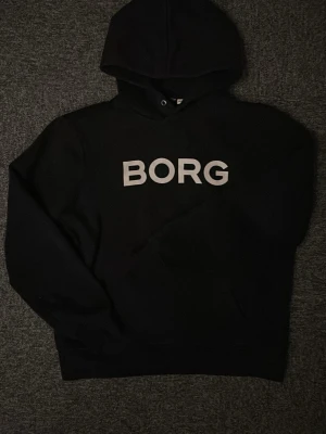 Svart hoodie från BORG  - Svart hoodie från BORG med stor vit logga på bröstet. Klassisk modell med huva och dragsko, samt ribbade muddar vid ärmslut och nederkant. Perfekt för en chill och sportig stil.
