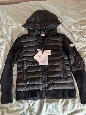 Svart dunjacka med huva från Moncler - Snygg svart dunjacka från Moncler med stickade ärmar och huva. Jackan har två fickor framtill med dragkedja och Moncler-logga på ena ärmen. Perfekt för kalla vinterdagar och riktigt stilren look.