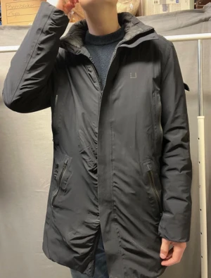 Svart parkas från Ubr - Snygg svart parkas från ubr med avtagbar huva och dragkedja framtill. Jackan har två snedställda fickor med dragkedja och diskret logga på bröstet. Perfekt för kalla dagar och enkel att matcha med olika outfits.