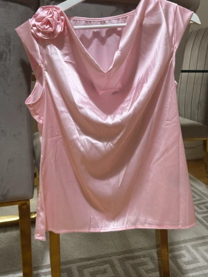 Ljusrosa satintopp med rosdetalj - Snygg ljusrosa topp i satin med draperad urringning och en dekorativ ros på axeln. Ärmlös modell som ger en elegant look och mjukt glansigt material som känns lyxigt. Perfekt för dig som vill sticka ut lite extra.
