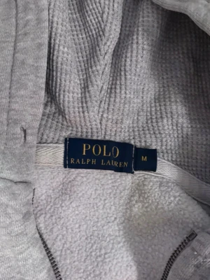 Grå hoodie från Polo Ralph Lauren - Snygg grå hoodie från Polo Ralph Lauren med klassisk blå logga broderad på bröstet. Mjuk insida och dragkedja framtill. Perfekt för en chill och stilren look. Materialet är bomullsmix som känns skönt mot huden. Huva med dragsko för extra komfort.