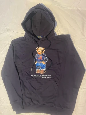 Navy hoodie Polo Bear Ralph Lauren - Navy hoodie från Ralph Lauren med den ikoniska Polo Bear-trycket framtill. Hoodien har huva med dragsko och en stor magficka. Materialet är mjukt och skönt, perfekt för en avslappnad stil. Klassisk passform och snygga detaljer.